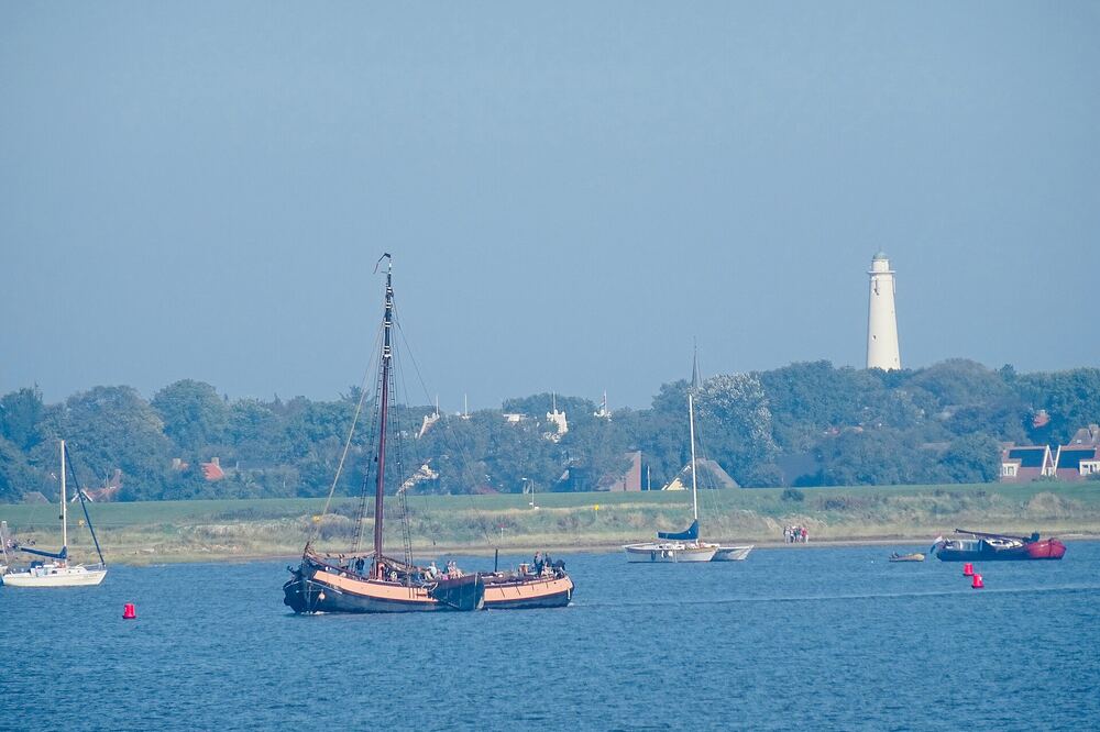 Zicht op Schiermonnikoog vanaf het water, met een zeilschip voor de kust en de vuurtoren boven de boomrand